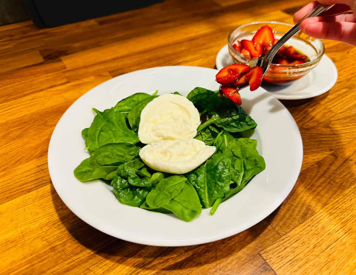 Sliced strawberries slicked with dressing being spooned onto spinach and burrata in a shallow white bowl.