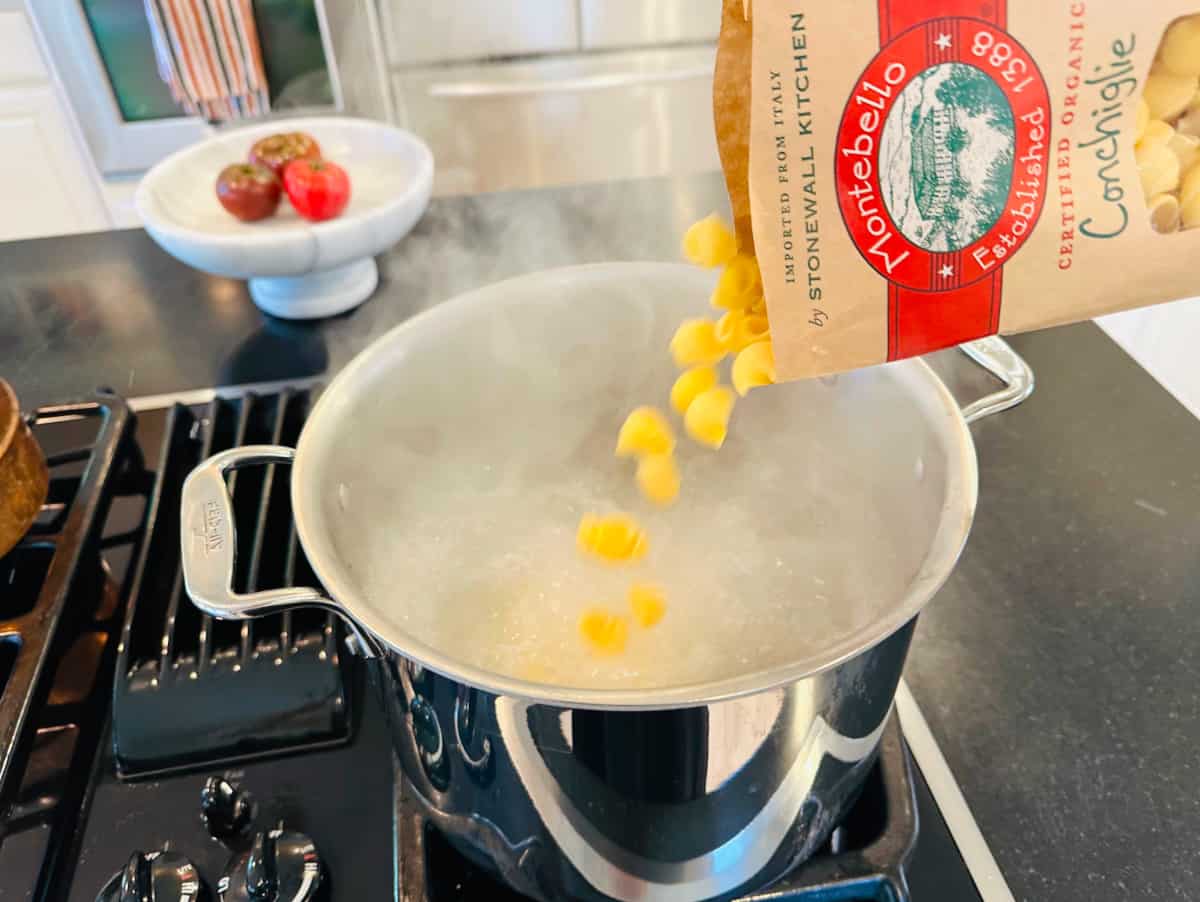 Shell shaped noodles being poured from a package into a large pot of boiling water on the stove.