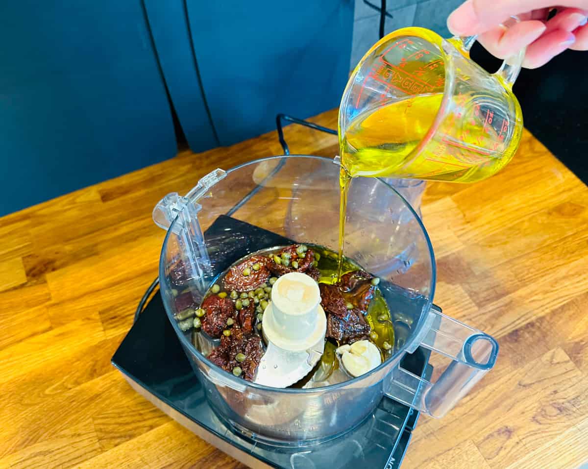 Olive oil being poured from a glass measuring cup into the bowl of a food processor with sun dried tomatoes, capers, and a clove of garlic in the bottom.