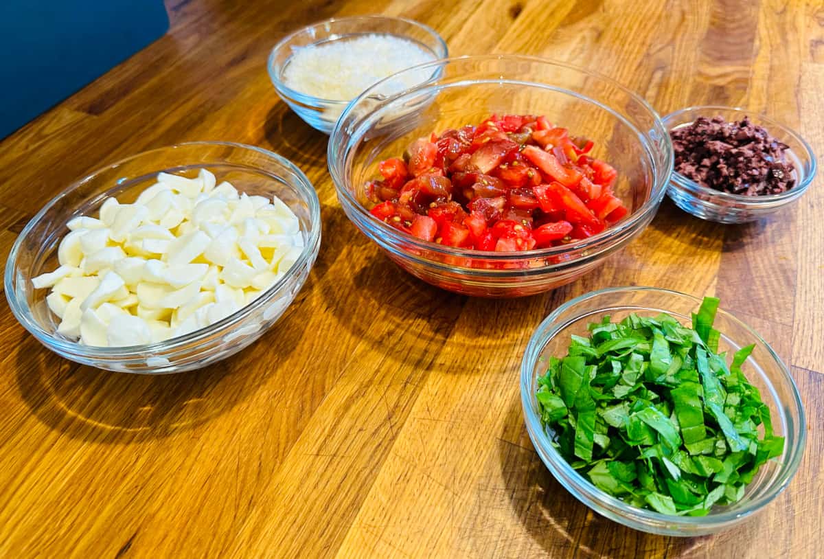 Separate glass bowls of chopped mozzarella, grated parmesan, chopped tomatoes, minced olives, and chopped basil.