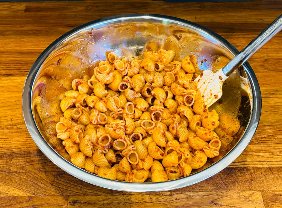 Shell shaped pasta coated in dressing in a large steel bowl with a white silicone spatula.