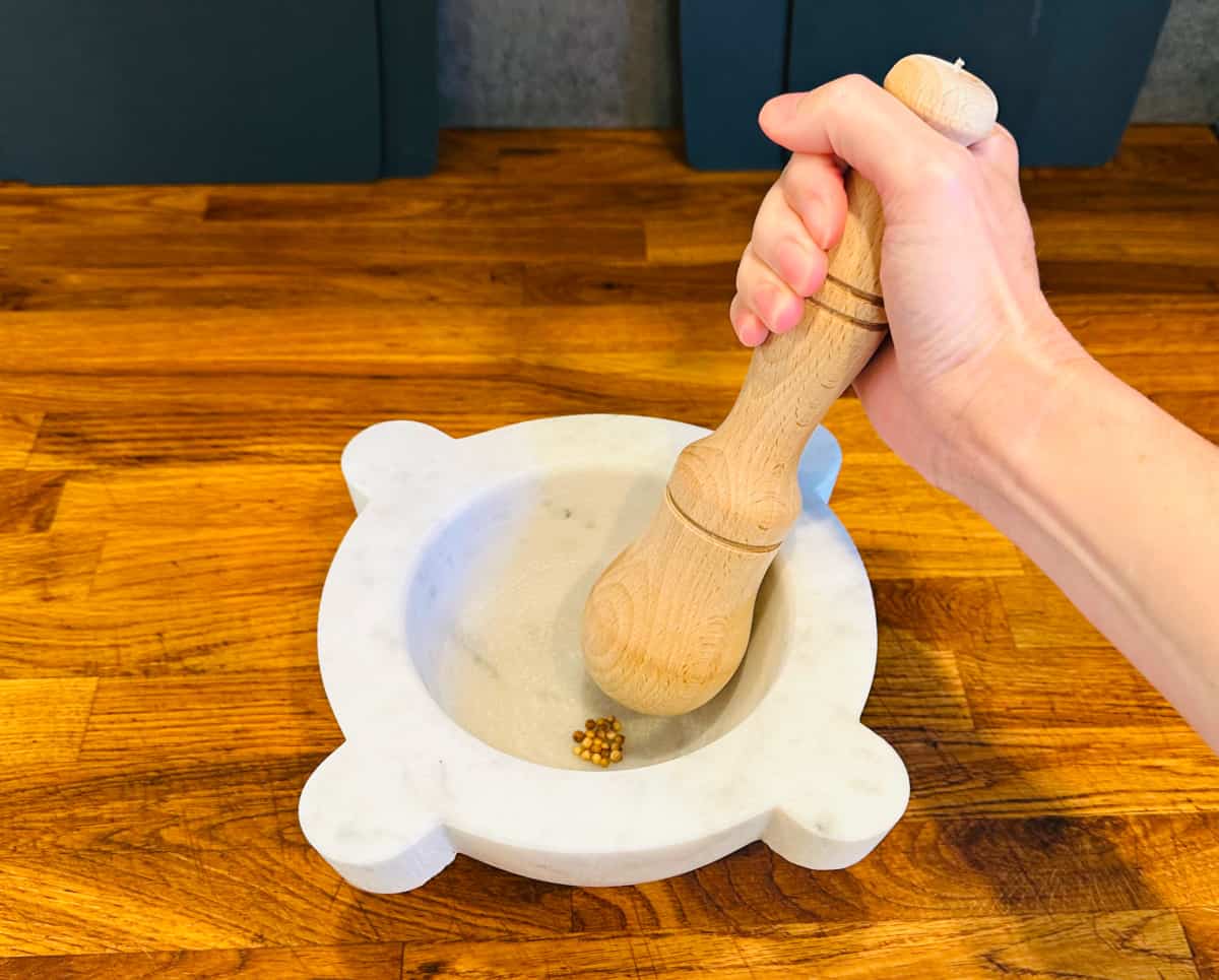 Small brown seeds being crushed with a large wooden pestle in a white marble mortar.