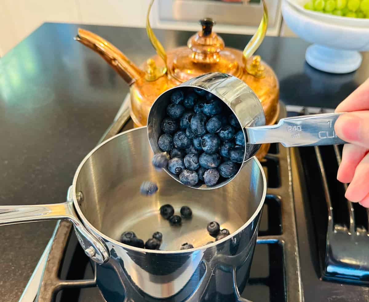 Blueberries being tipped out from a steel measuring cup into a small steel saucepan on the stove.