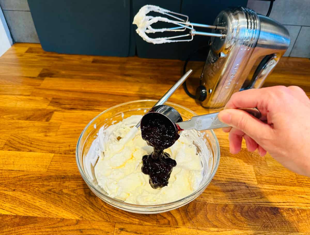 Dark berry mixture being poured from a steel measuring cup into whipped cream in a glass bowl next to a silver electric mixer.