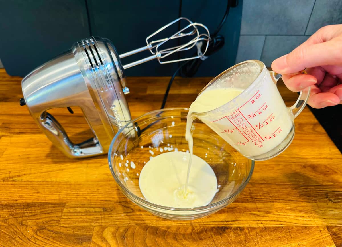 Whipped cream being poured from a glass measuring cup into a glass bowl sitting next to a silver electric mixer.
