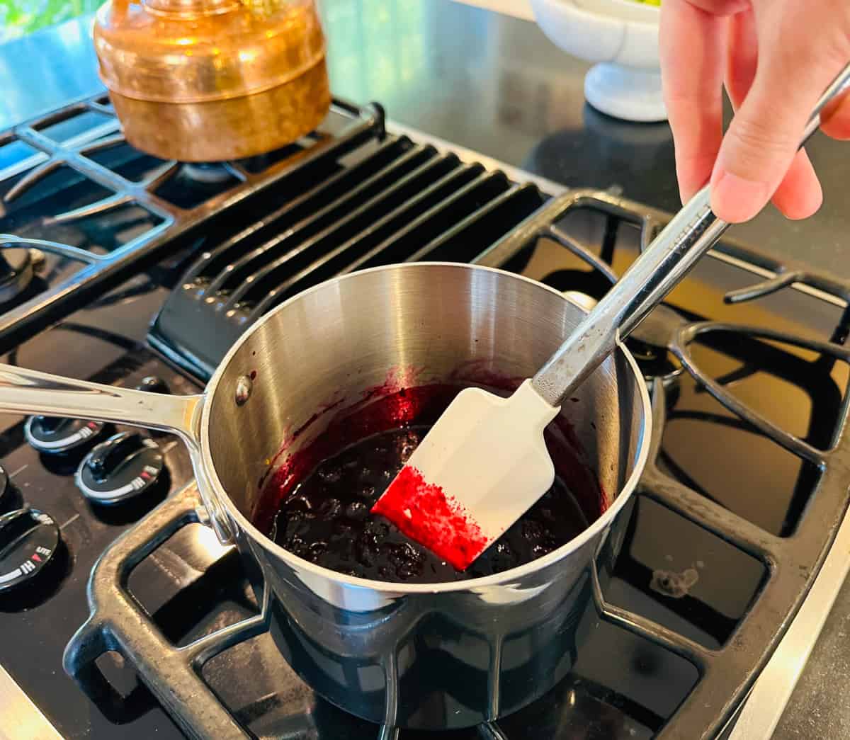 Blueberry compote being stirred with a white silicone spatula while cooling in a small steel saucepan.