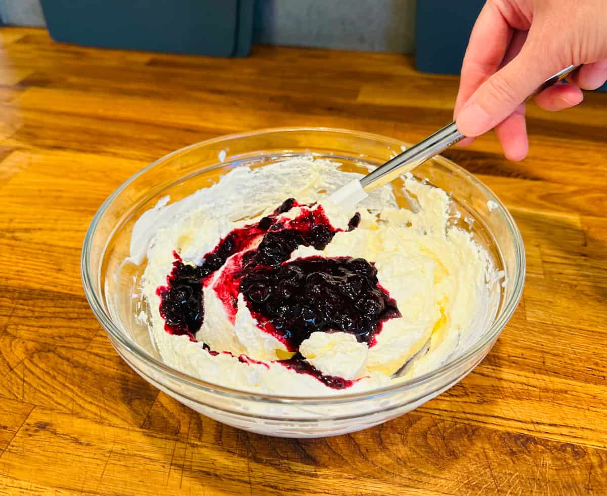 Blueberry compote being folded by a white silicone spatula into whipped cream in a glass bowl.