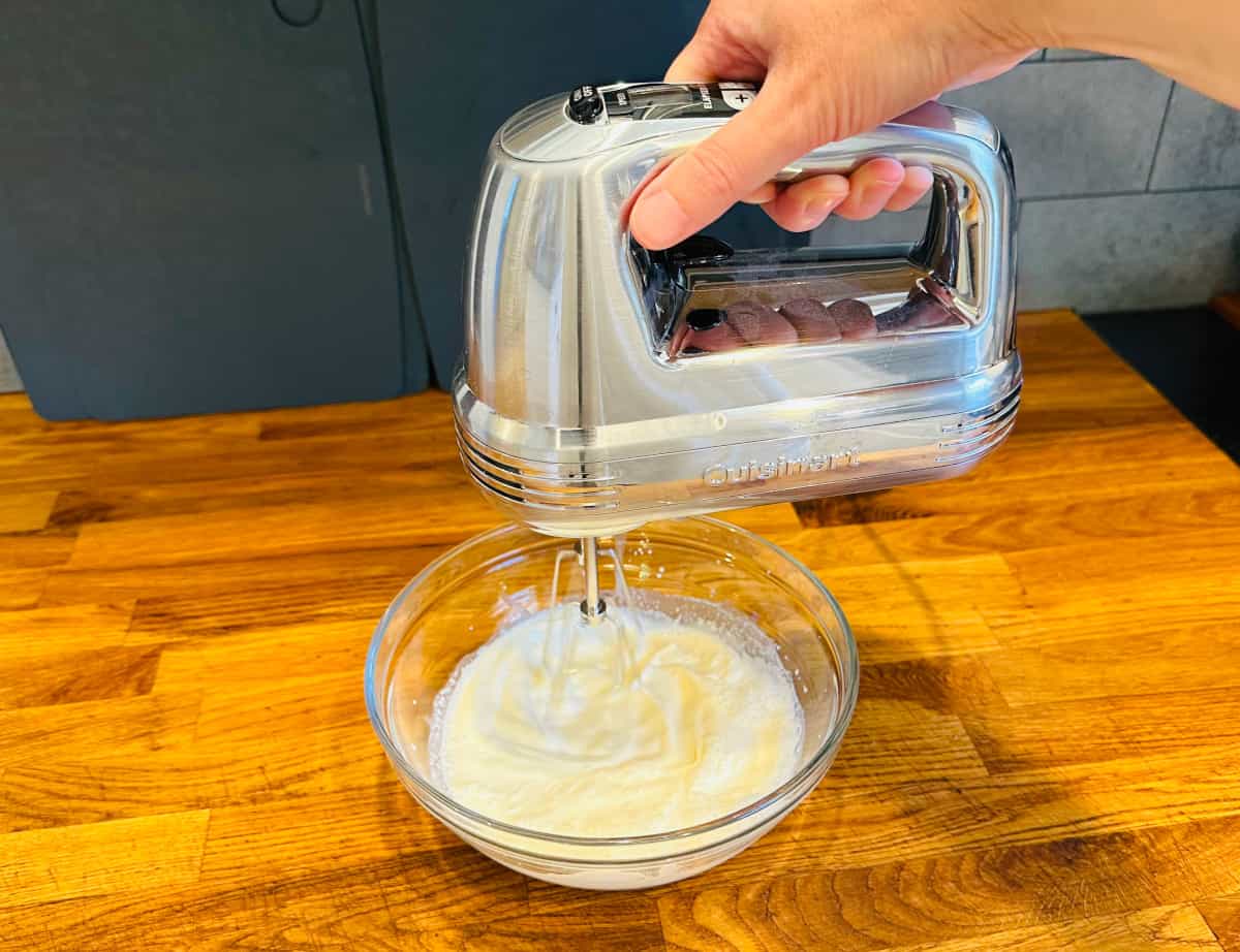 Cream being whipped with a silver electric mixer in a glass bowl.