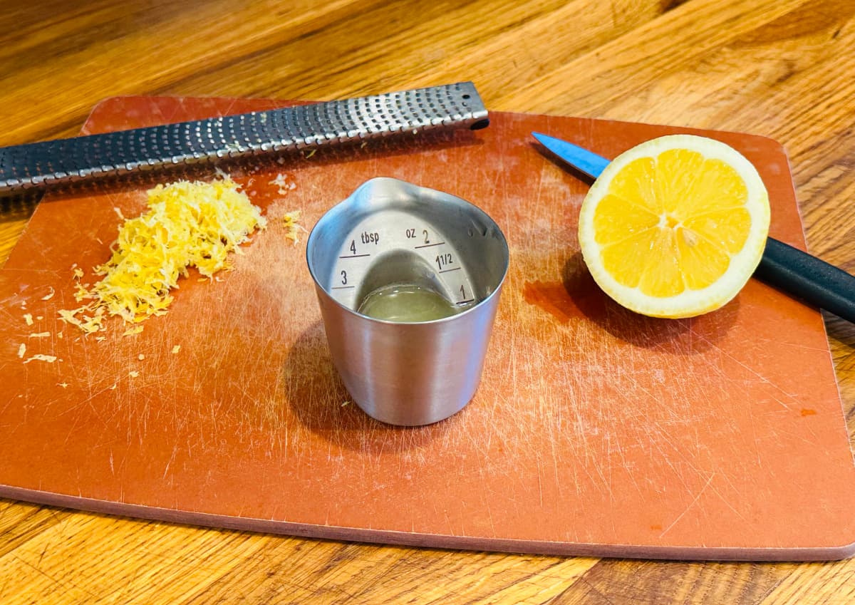 Lemon zest, lemon juice in a steel measuring jigger, and half a lemon on a cutting board with a zester and small knife.