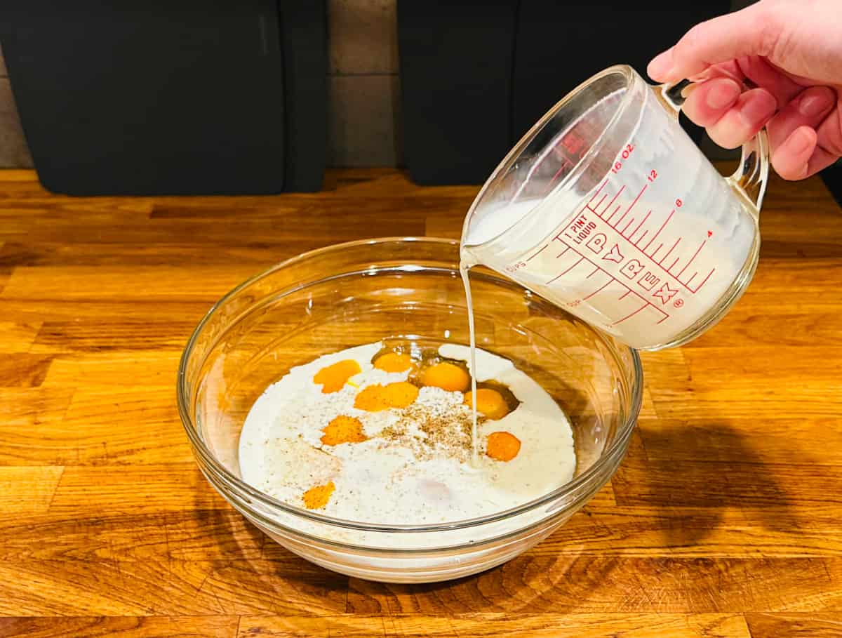 Half and half being poured from a glass measuring cup into a glass bowl full of raw eggs.
