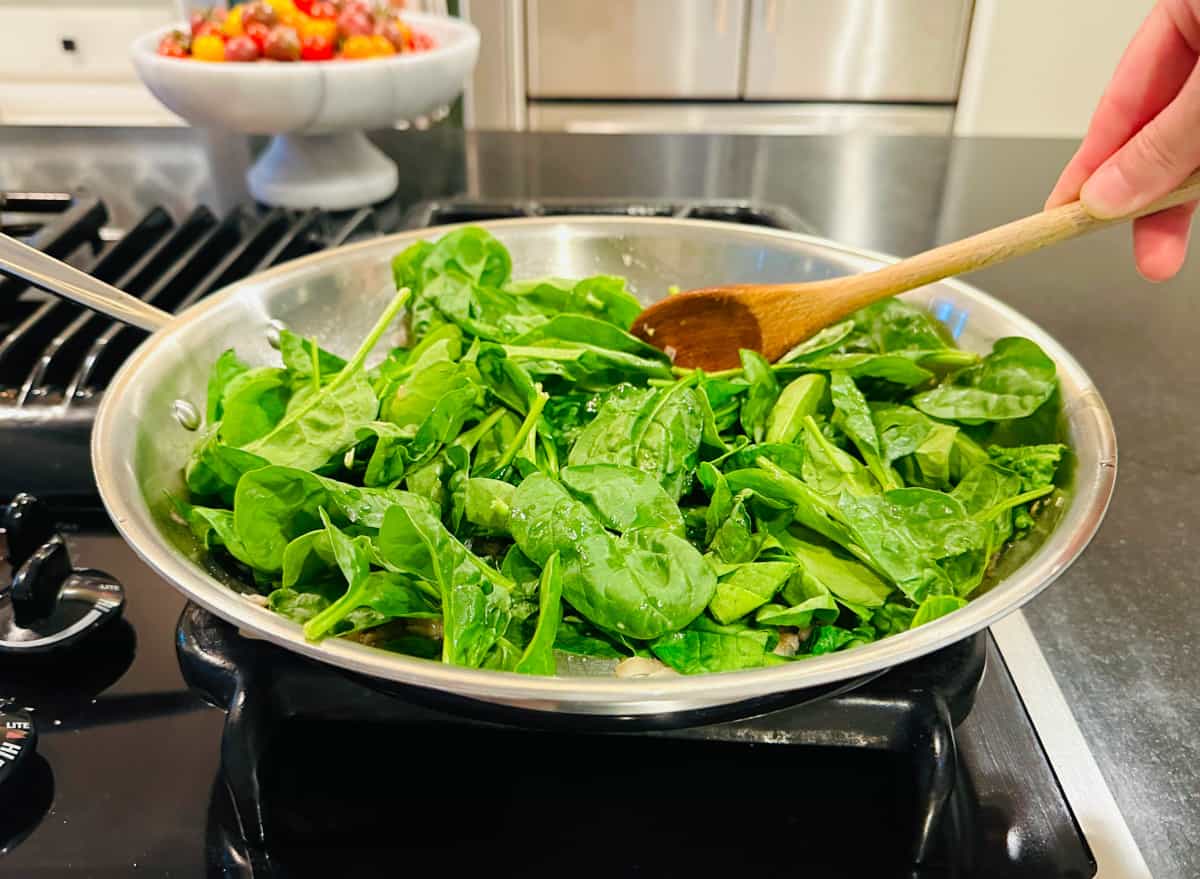 Baby spinach being stirred with a wooden spoon in a large steel skillet on the stove.
