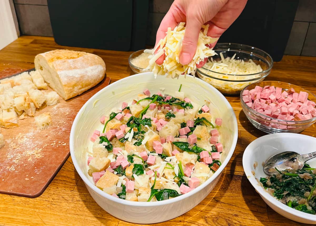 Shredded white cheese being sprinkled over cubes of bread, chunks of ham, and cooked spinach in an oval white baking dish.