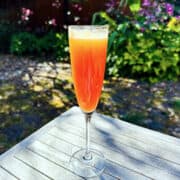 Breakfast at Persephone's (AKA Peach & Pomegranate Bellini) in a champagne flute sitting on a wooden outdoor table surrounded by some shrubs and flowering plants.