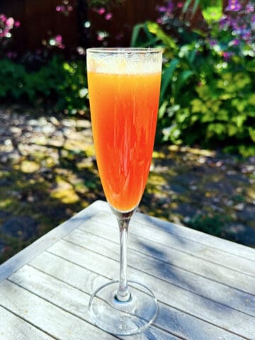 Breakfast at Persephone's (AKA Peach & Pomegranate Bellini) in a champagne flute sitting on a wooden outdoor table surrounded by some shrubs and flowering plants.