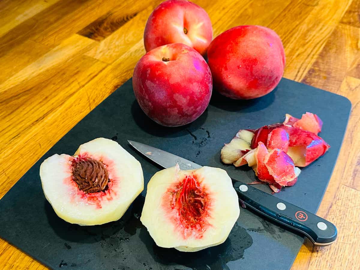 Two halves of a white peach sitting next to a small knife, a pile of peach peels, and three whole peaches on a dark gray cutting board.