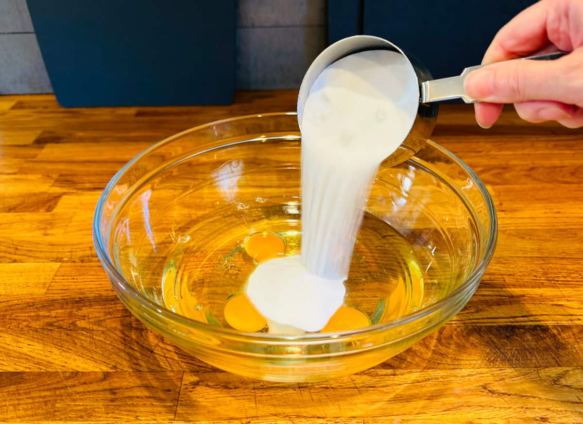 Sugar being poured from a steel measuring cup into a glass bowl containing eggs and vegetable oil.
