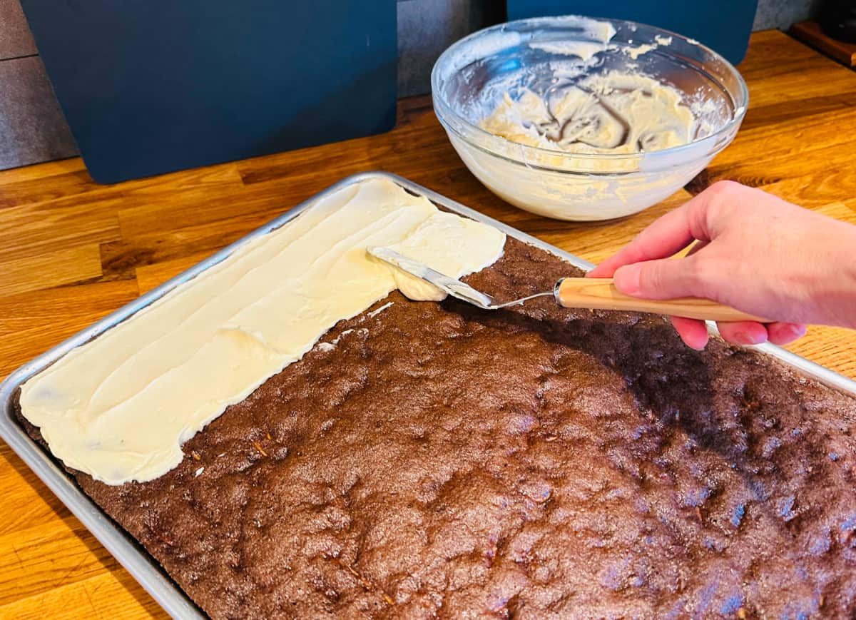 Cream cheese frosting being spread over a pan of chocolate zucchini bars with an offset spatula.