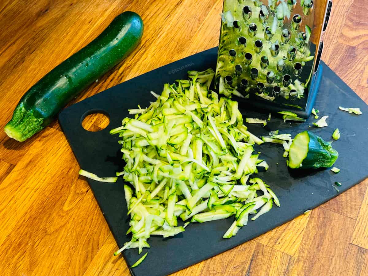 A pile of shredded zucchini next to a box grater on a dark gray cutting board.