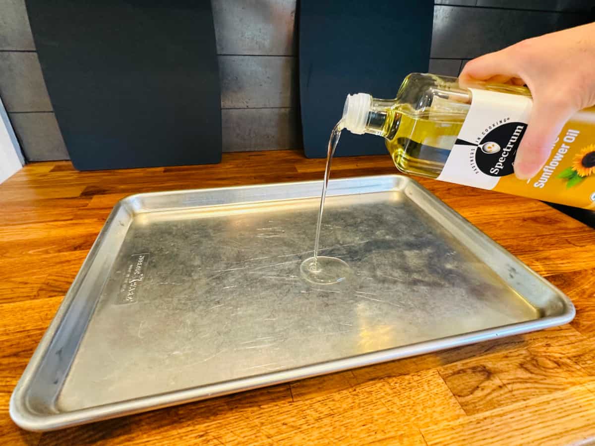 Pale yellow liquid being poured from a bottle of sunflower oil into a metal jelly roll pan.