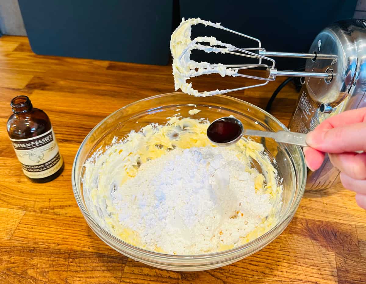 Dark brown liquid in a steel measuring spoon being held over a glass bowl containing powdered sugar, butter, and cream cheese next to a bottle of vanilla extract and an electric mixer.