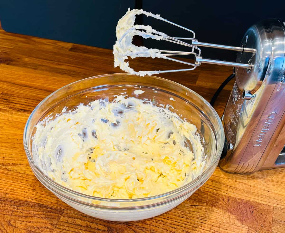 Pale yellow mixture of butter and cream cheese in a glass bowl next to an electric mixer.