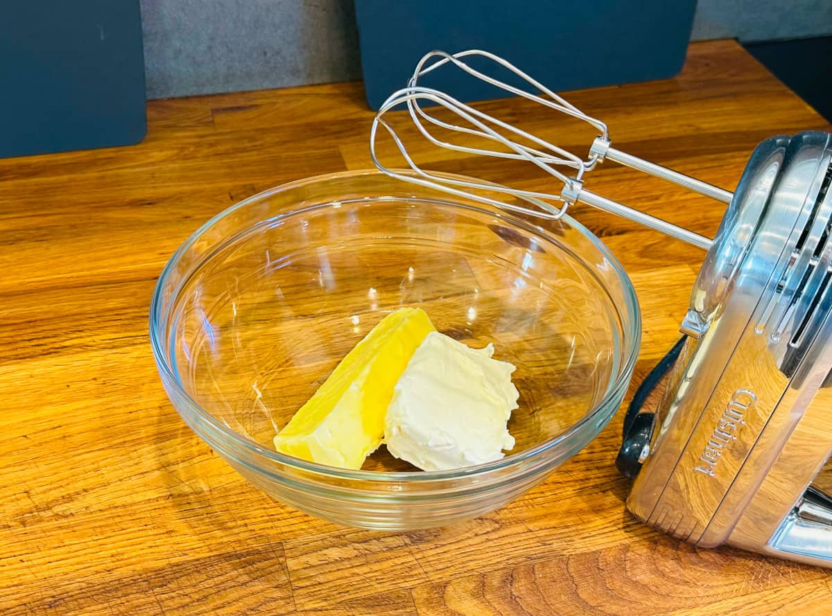 A glass bowl containing a cube of butter and block of cream cheese next to an electric mixer.