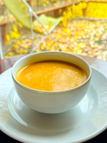 Orange soup in a white bowl sitting on a dark wood table in front of a window looking out on a lawn chair sitting on a deck covered in yellow leaves.