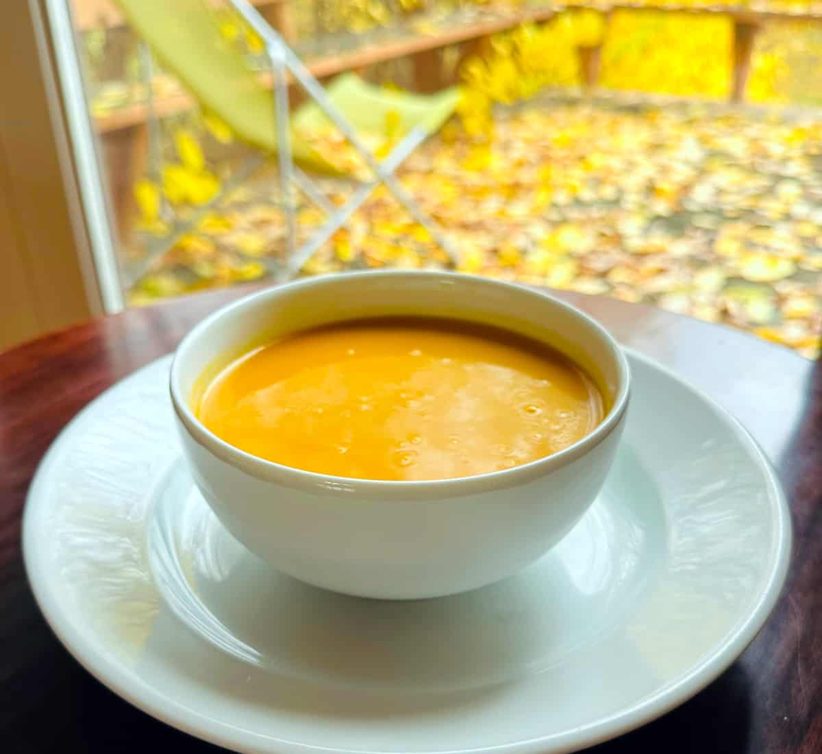 Creamy carrot soup in a white bowl sitting on a dark wood table in front of a window looking out on a lawn chair sitting on a deck covered in yellow leaves.