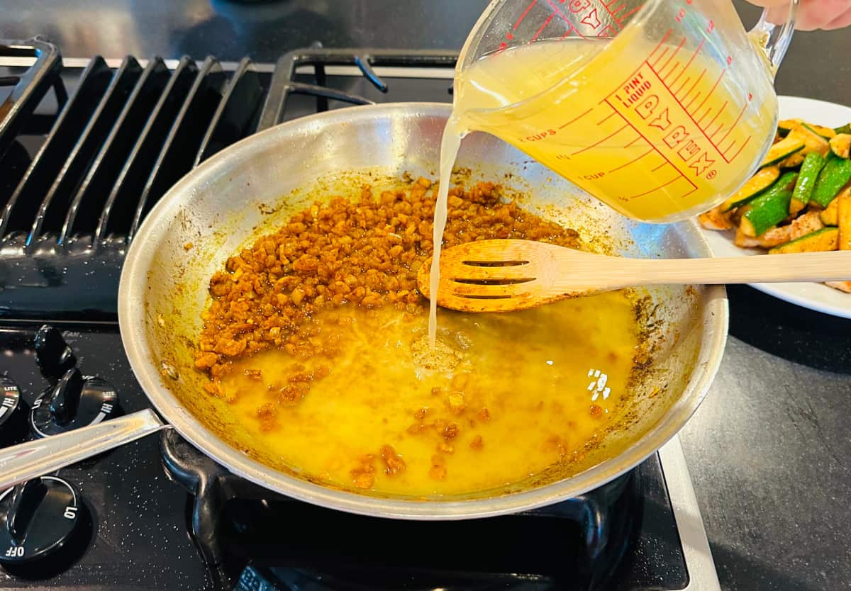 Chicken broth being poured from a glass measuring cup into a large steel skillet containing browned onions and a wooden slotted spoon.