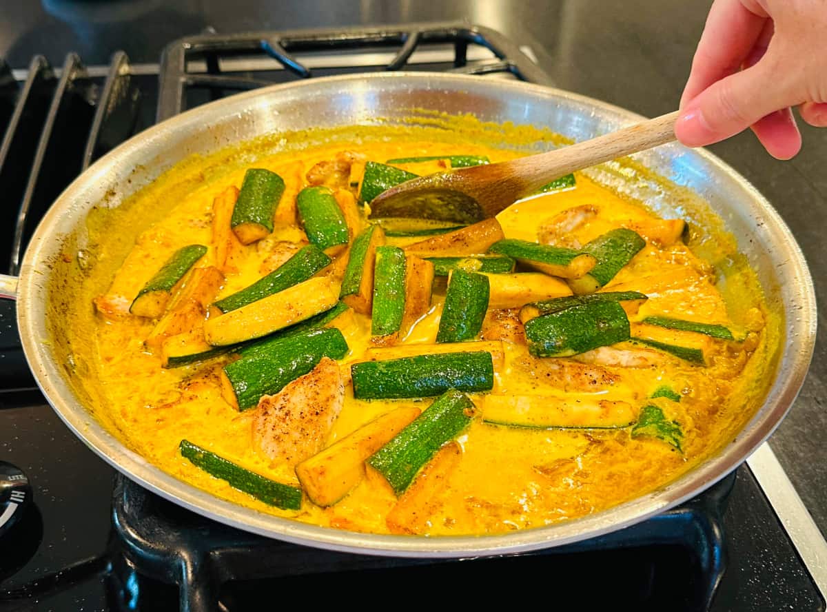 Curried chicken and zucchini being stirred with a wooden spoon in a large steel skillet on the stove.