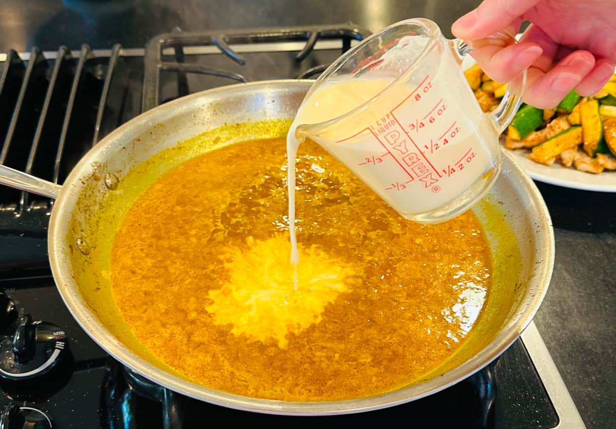 Cream being poured from a glass measuring cup into a large steel skillet containing brown liquid.