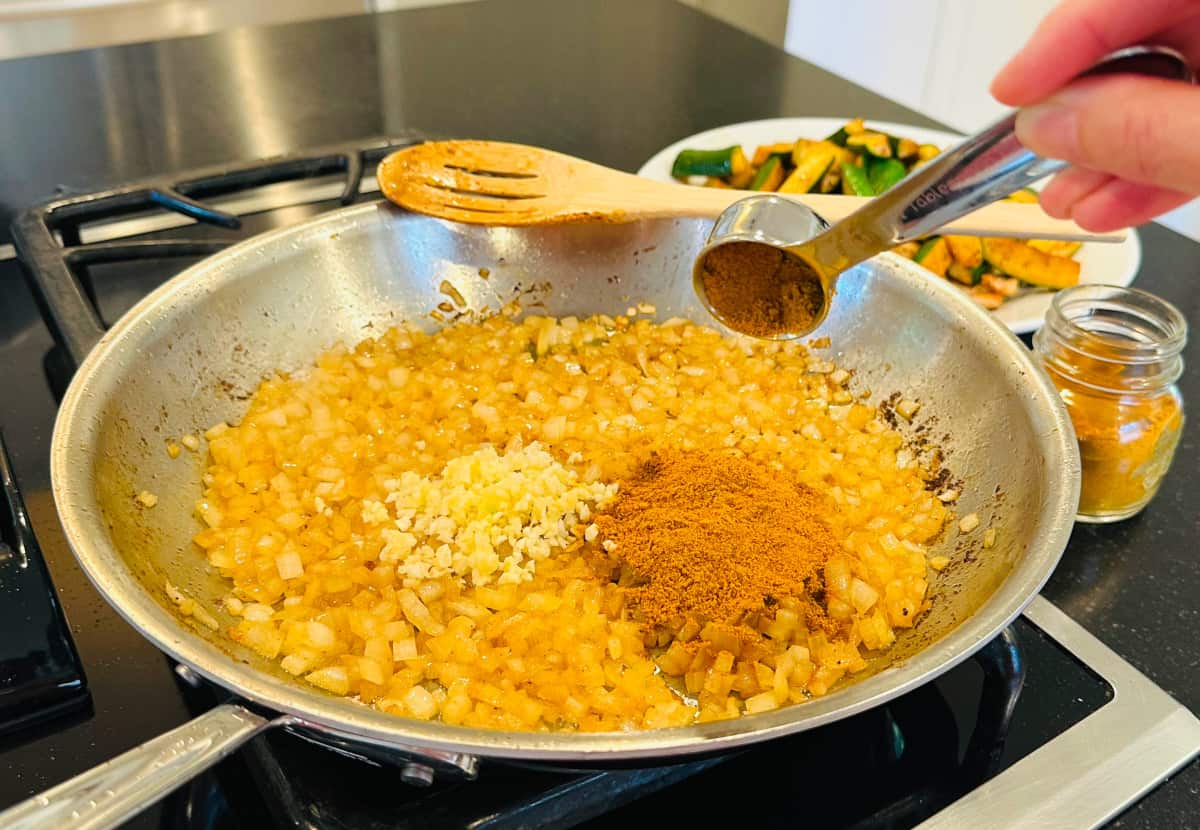 Yellow curry powder being tipped from a steel measuring spoon into a large steel skillet containing browned onions and a small heap of minced garlic.