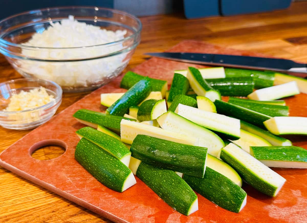 Zucchini spears on a cutting board next to a medium glass bowl of chopped onions and a small glass bowl of minced garlic.