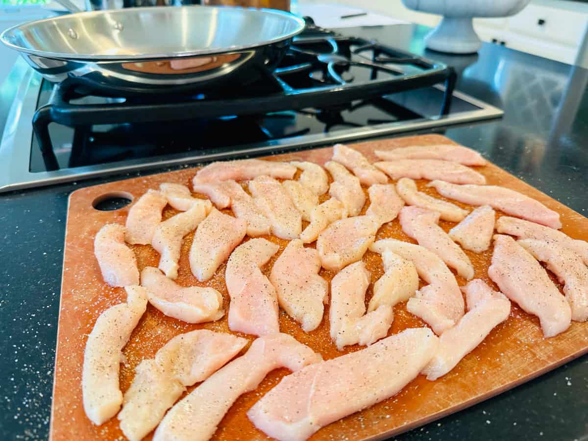 Raw slices of chicken on a cutting board next to a large steel skillet sitting on a stove.