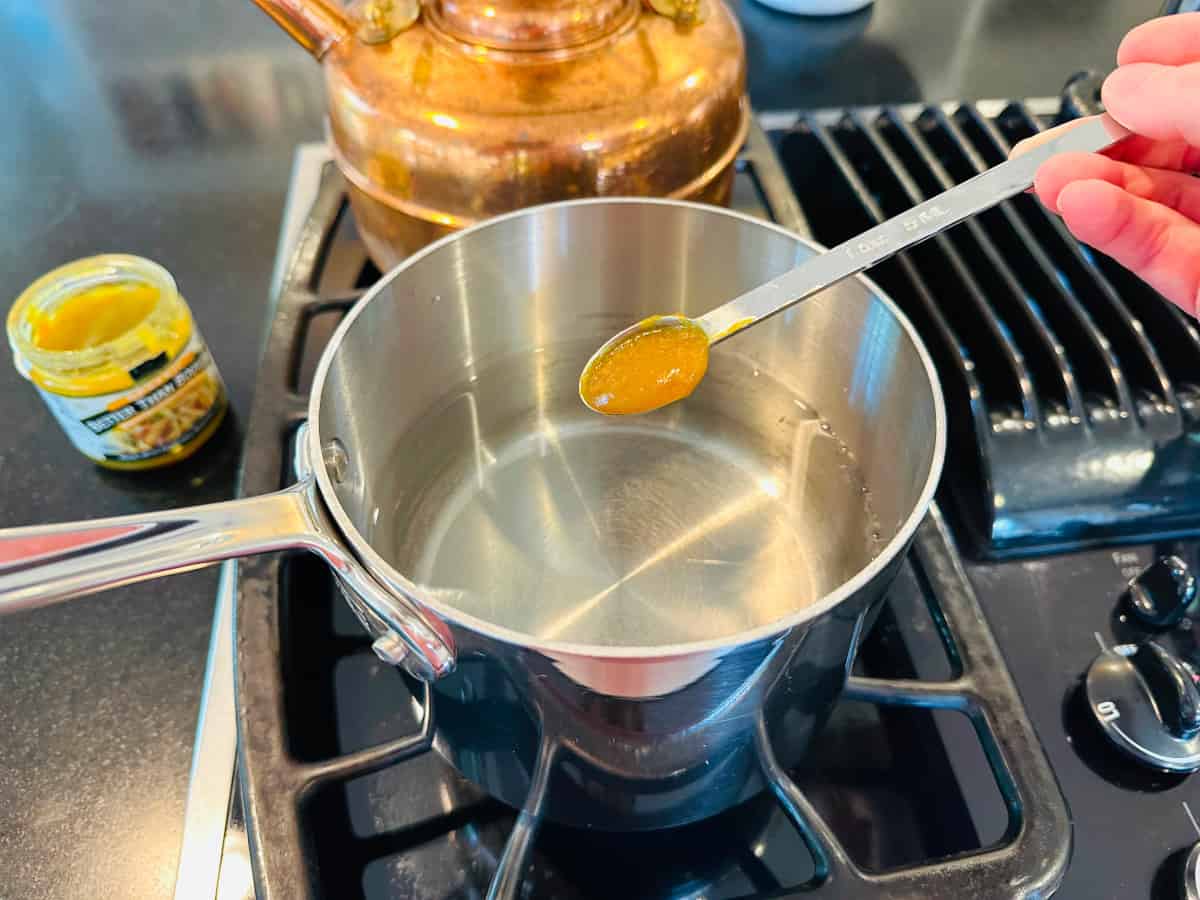 Chicken bouillon concentrate in a steel measuring spoon being held over a small steel saucepan of water on the stove.
