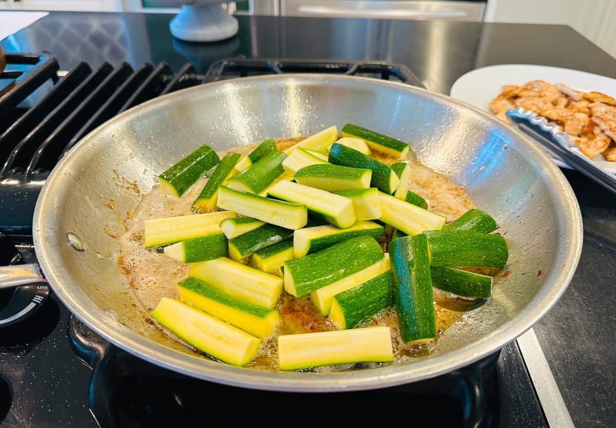 Zucchini spears cooking in a large steel skillet on the stove.