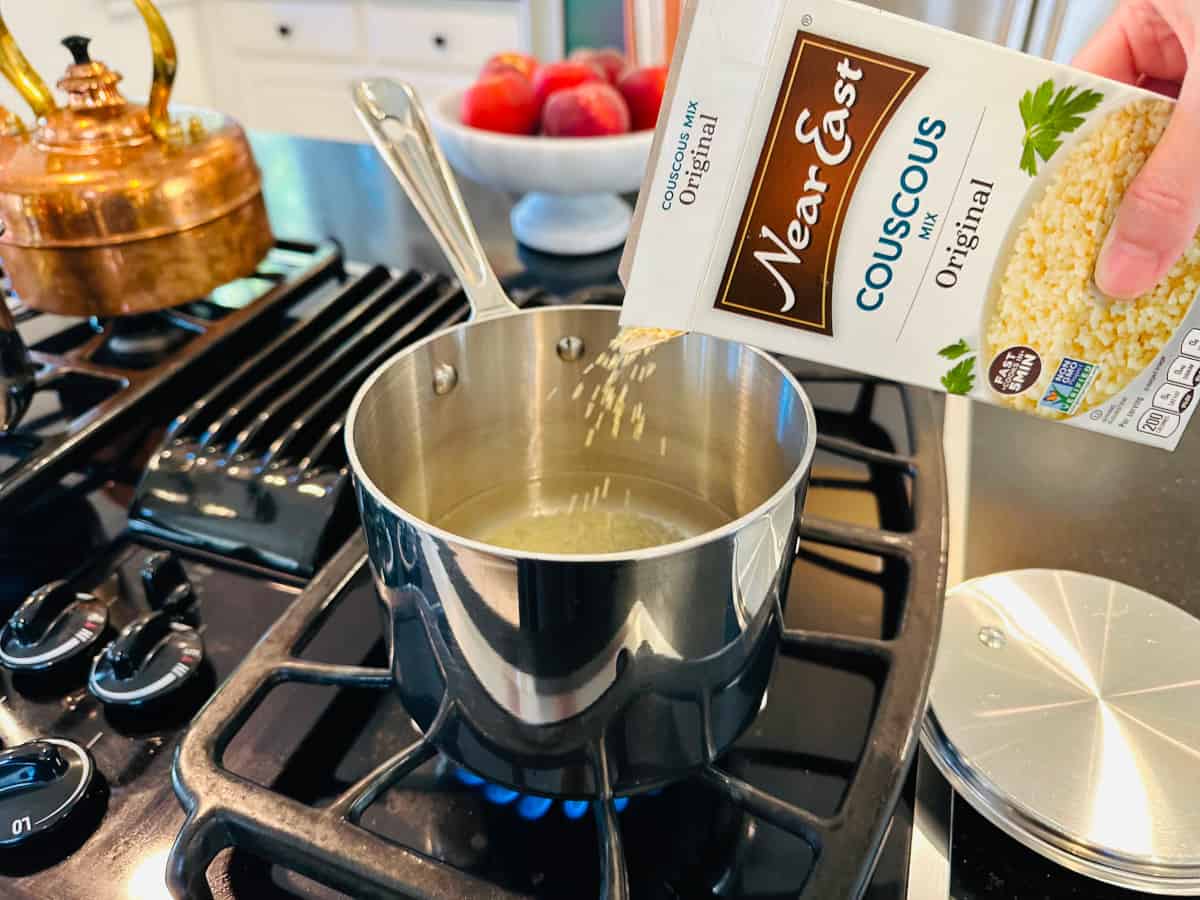 Couscous being poured from a box into a small steel saucepan of boiling water on the stove.