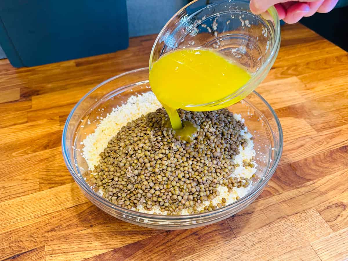 Yellow colored dressing being poured from a small glass bowl into lentils and couscous in a large glass bowl.