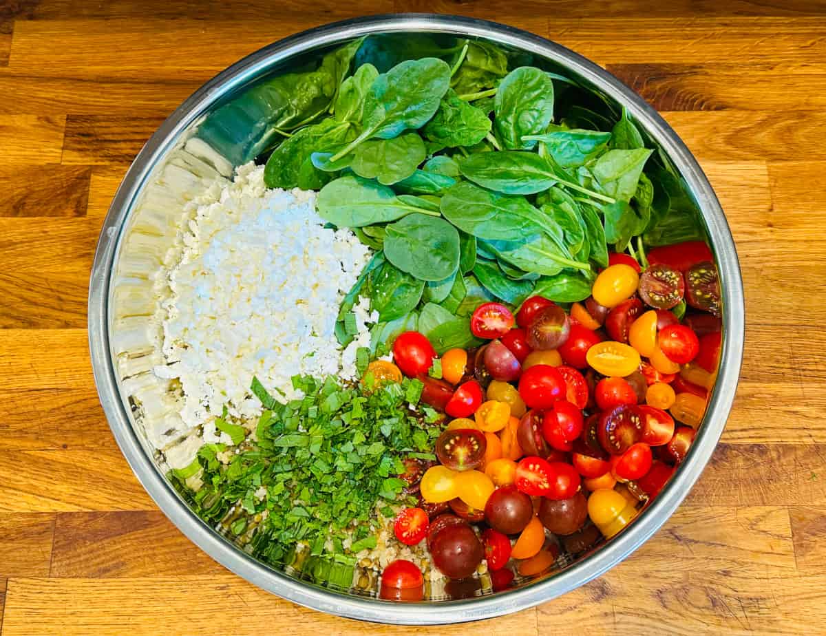 Distinct heaps of baby spinach, crumbled feta, chopped mint, and cherry tomatoes in a large steel bowl.