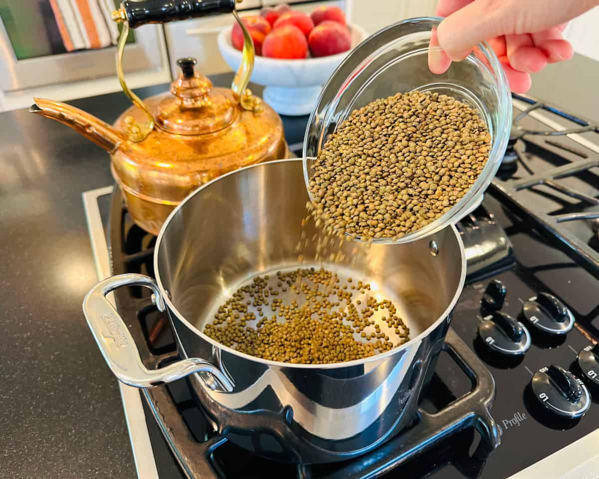 Green lentils being tipped out from a glass bowl into a medium steel saucepan.