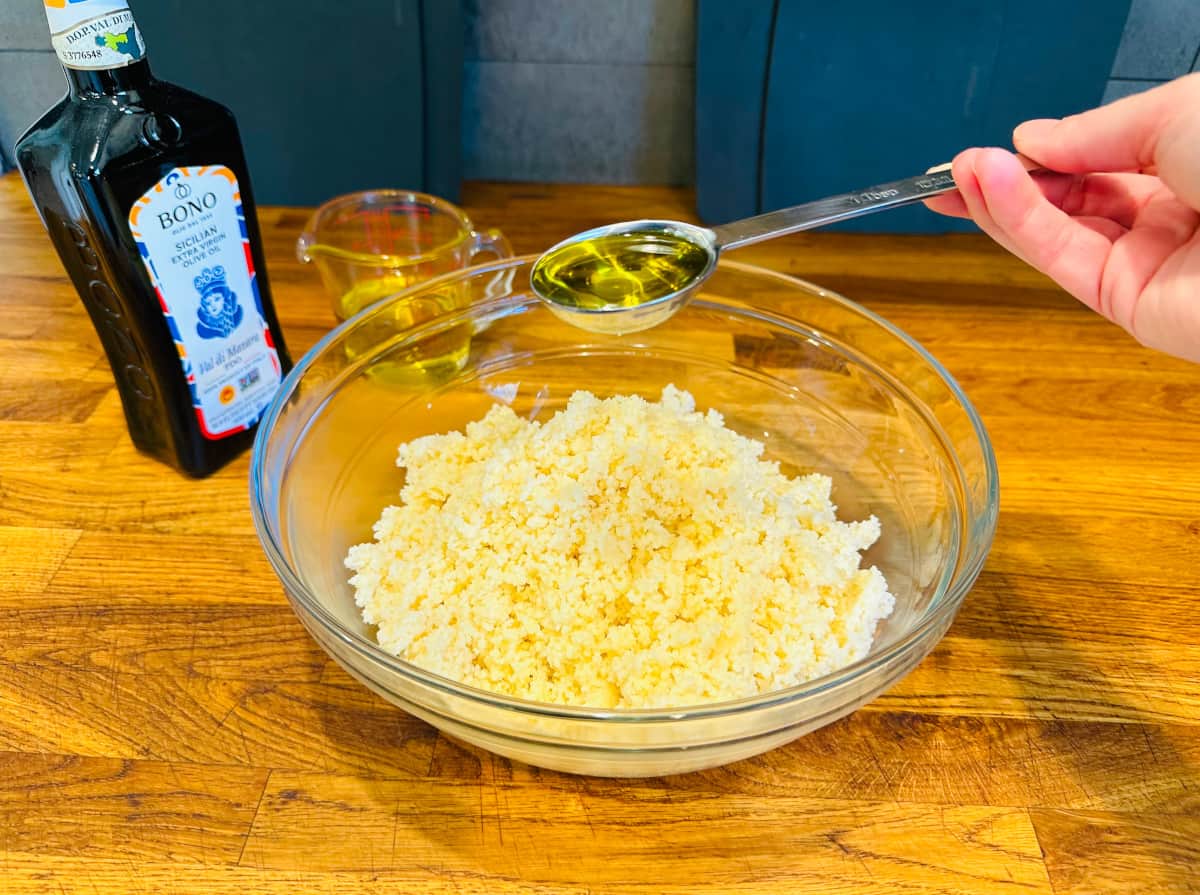 Steel measuring spoon filled with olive oil being held over a large glass bowl of couscous.