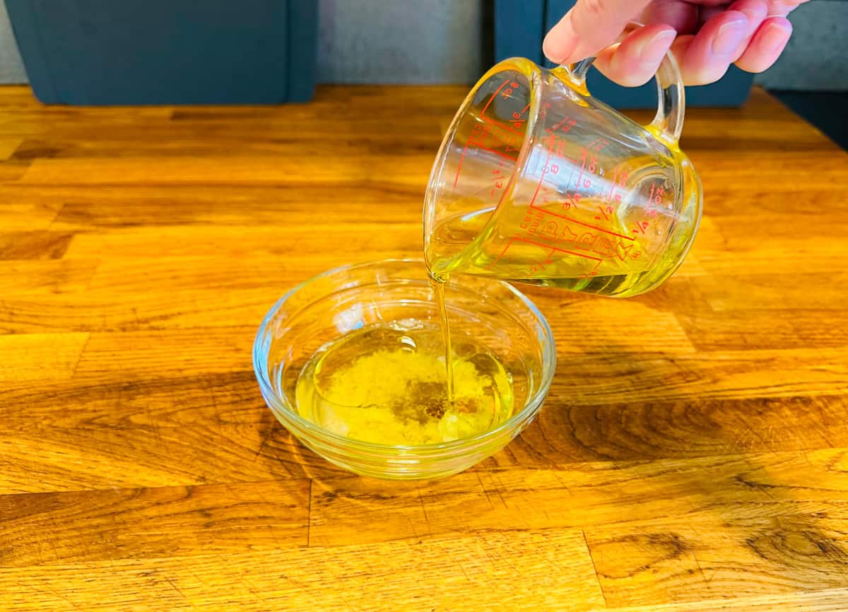 Olive oil being poured from a glass measuring cup into a small glass bowl containing white vinegar and grated garlic.