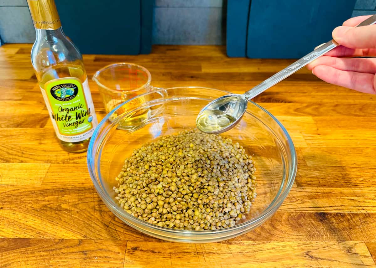 White vinegar being poured from a steel measuring spoon into a glass bowl of cooked lentils.