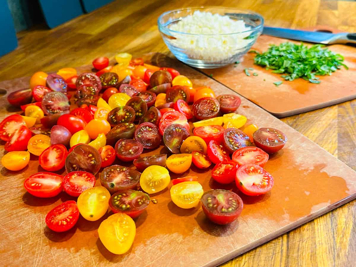 Halved red, orange, and yellow cherry tomatoes on a cutting board in front of chopped mint and a small glass bowl of crumbled feta.