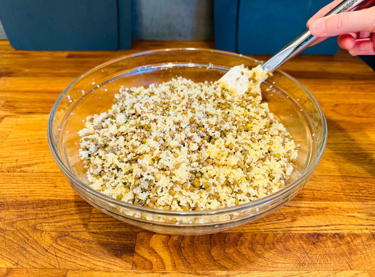 Couscous and lentils being stirred with a white silicone spatula in a large glass bowl.