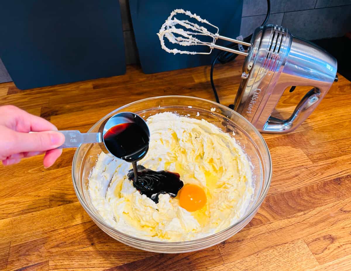 Molasses being poured from a steel measuring cup into a glass bowl containing creamed butter and sugar with an egg next to an electric mixer.