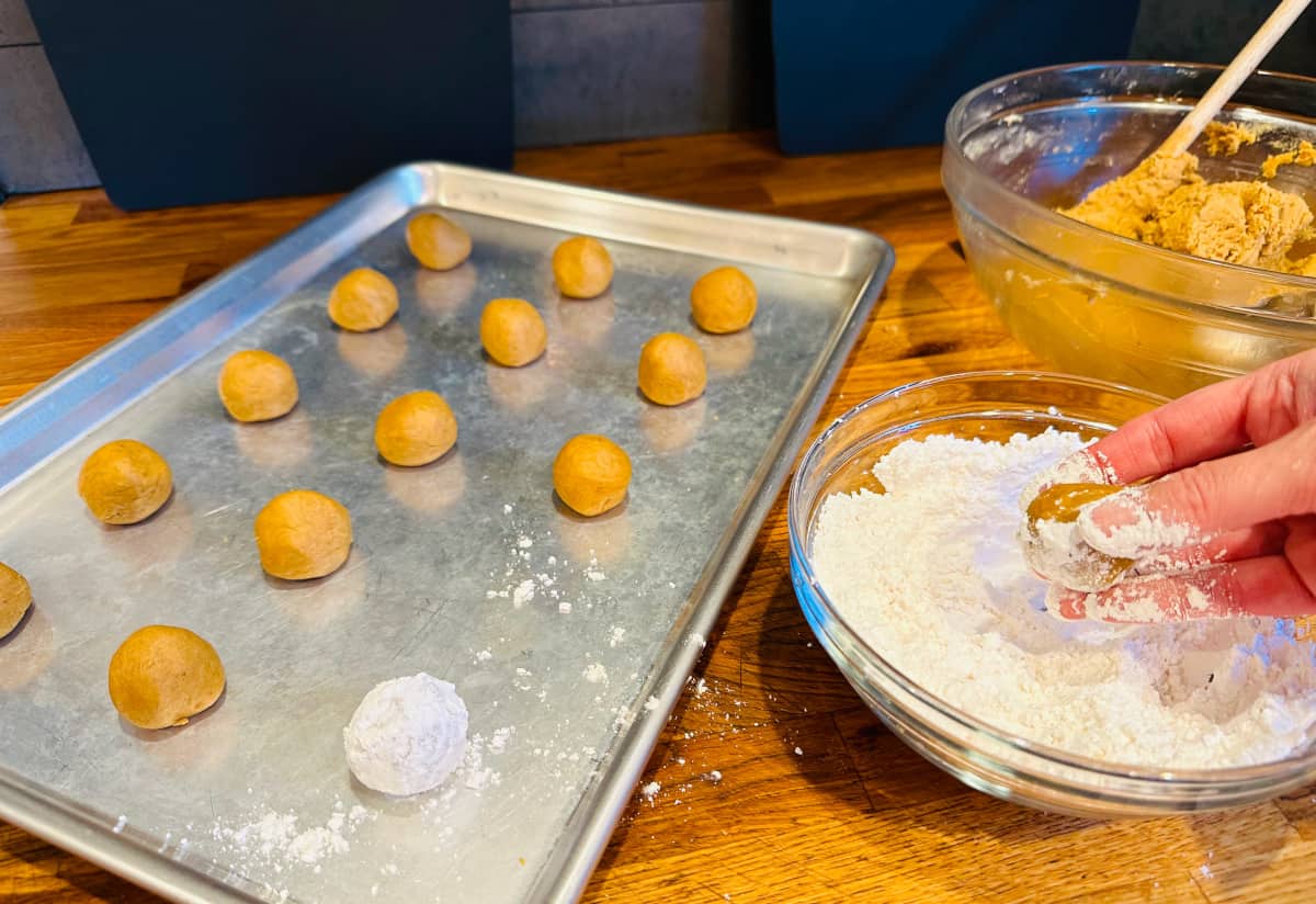 A ball of cookie dough being dipped in a small glass bowl of powdered sugar next to a metal baking sheet filled with small brown balls of cookie dough and a glass bowl of cookie dough.
