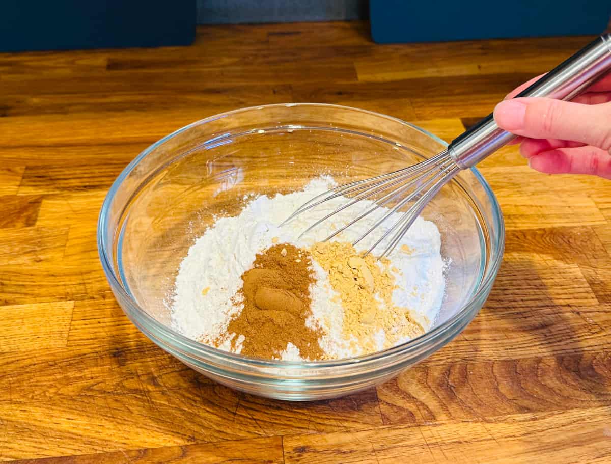 Flour, cinnamon, and ginger being stirred with a metal whisk in a glass bowl.