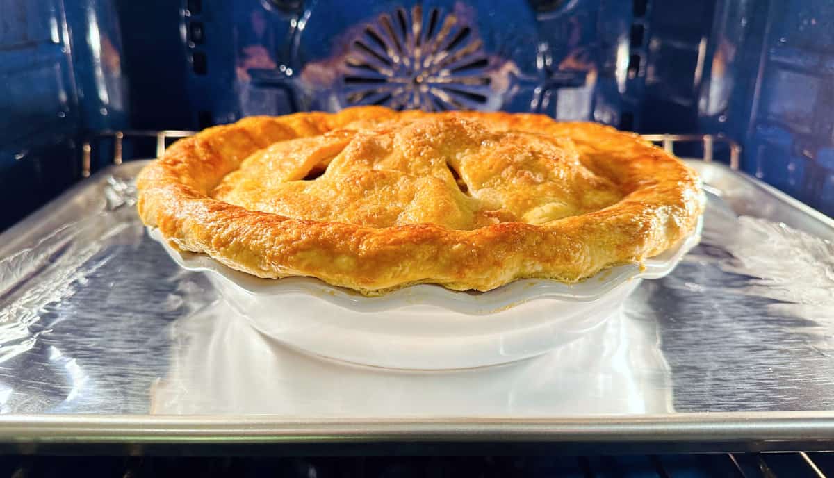 Old fashioned apple pie in a white ceramic pie plate sitting on a metal baking sheet in a blue oven.