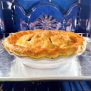 Old fashioned apple pie in a white ceramic pie plate sitting on a metal baking sheet in a blue oven.